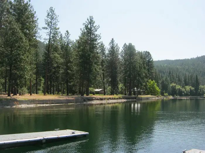 The photo shows a calm lakeshore lined with tall pines and a small dock in the foreground, with forested hills rising in the background